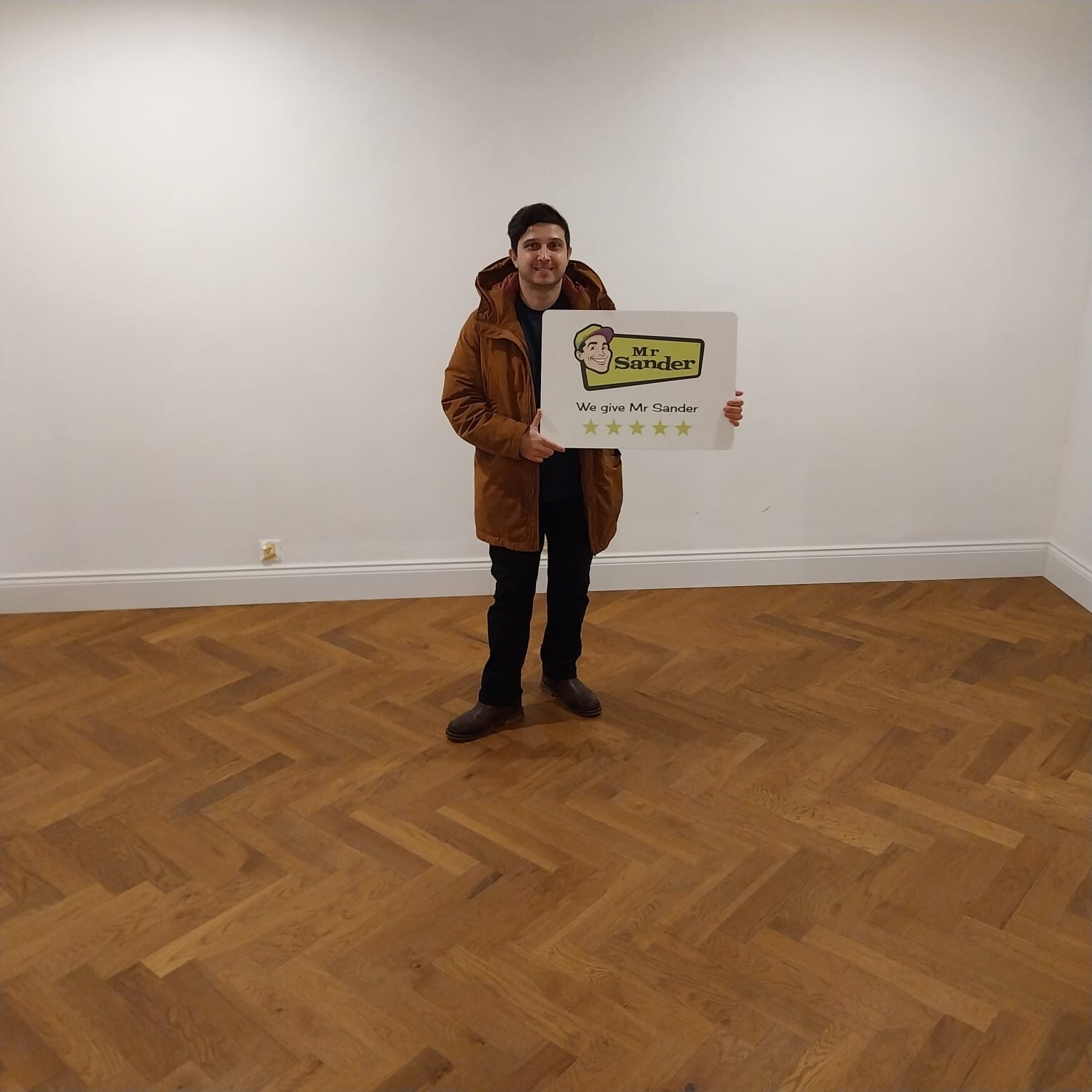 Happy customer standing on a newly restored herringbone oak parquet wood floor after professional Parquet Floor Refitting, holding a Mr Sander® five-star review sign.