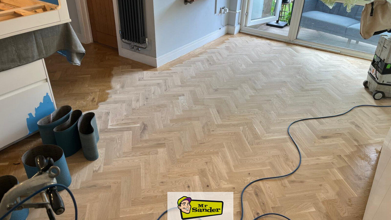 Parquet Floor Restoration — herringbone oak floor mid-sanding in a kitchen/diner, showing the contrast between the darker unsanded area and the pale, freshly sanded section; sanding belts and dust-extraction hose visible.