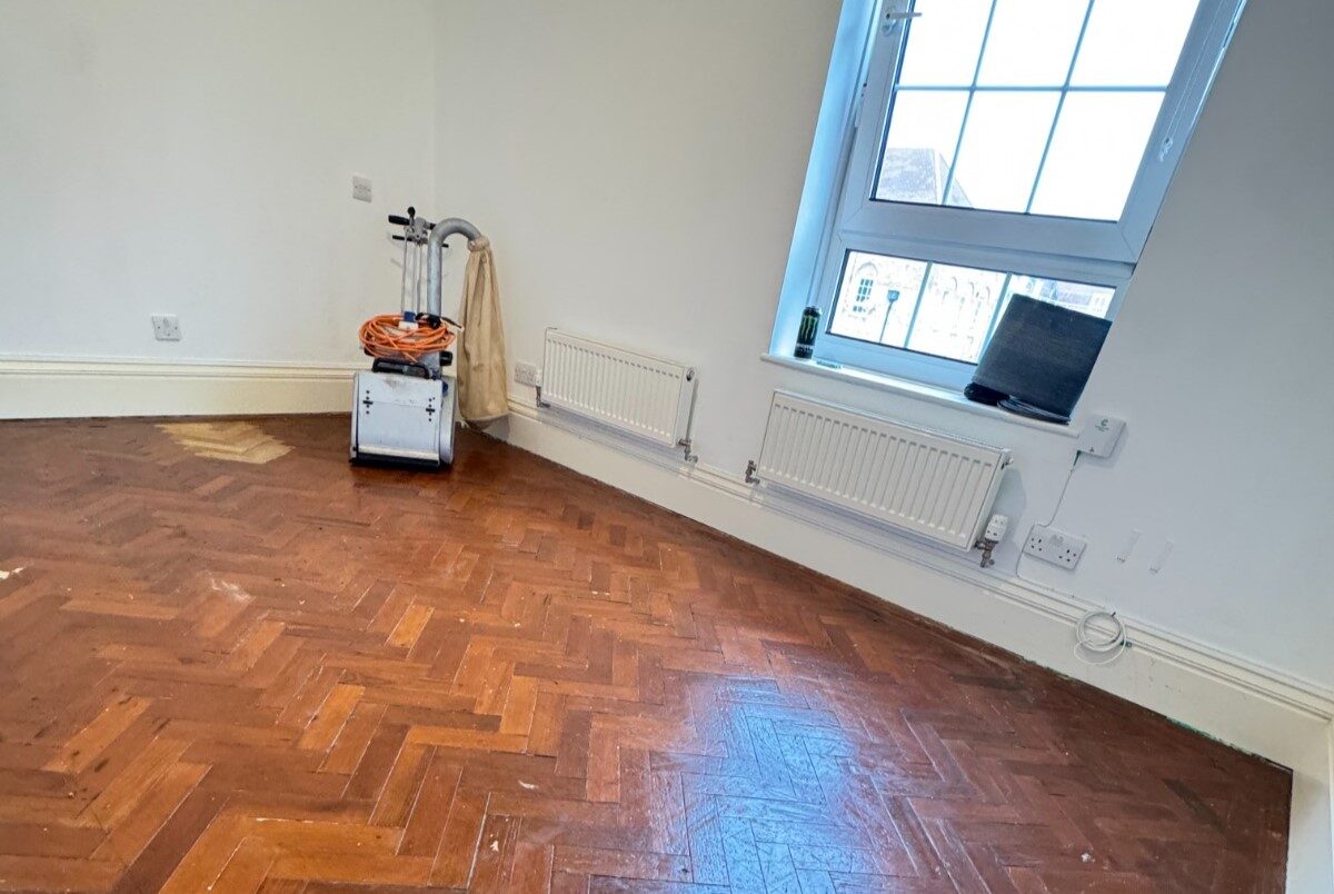 Parquet Floor Sanding preparation on a worn herringbone parquet wood floor with a sanding machine positioned in the corner of a bright room