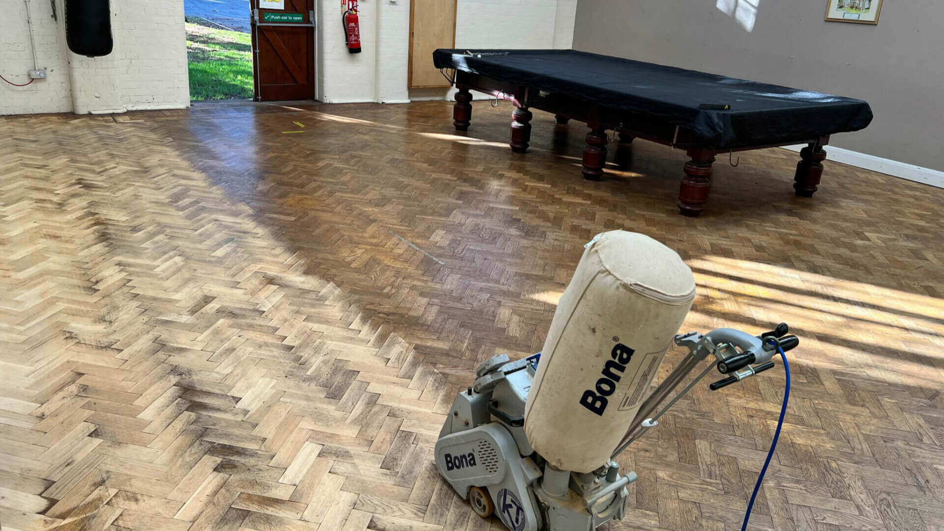 Parquet Floor Sanding in a large hall with an oak herringbone parquet wood floor, showing a Bona sanding machine and a partially sanded surface around a games table