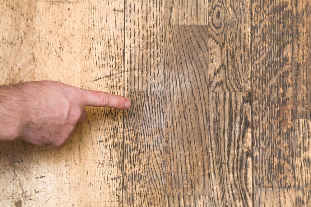 Close-up of a finger indicating a deep scratch in worn oak floorboards that show contrasting light and dark patches.