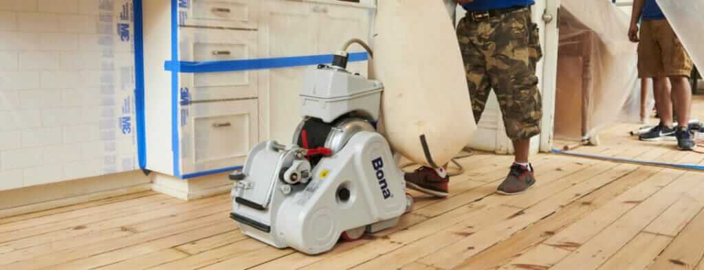 Professional man from Mr Sander® using Bona Belt UX (AMK173100) belt sander to sand pine floor in, UK living room. Specifications: 2.2 kW effect, 230V voltage, 50Hz frequency, 200x750mm size, 200mm drum width, 200x750mm sanding belt size, 2.2 kW motor power, 85kg weight.
