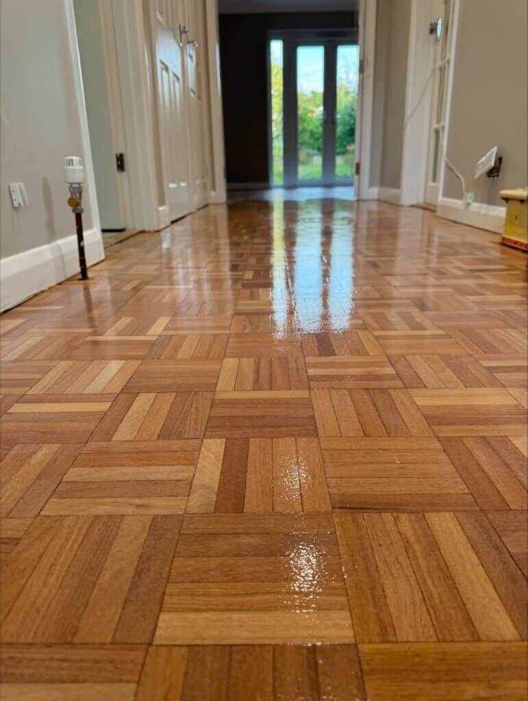 A freshly sanded and refinished parquet wood floor in a hallway, featuring a square geometric pattern. The glossy finish beautifully reflects the natural light coming from a glass door at the end of the hallway, enhancing the intricate detail and warmth of the parquet flooring. The polished floor contrasts elegantly with the neutral-toned walls and white doors, adding to the sophisticated look of the space.