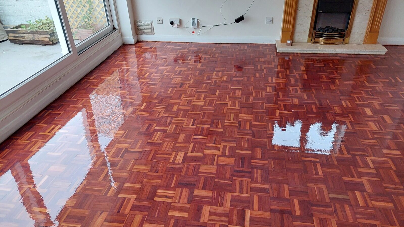 High-gloss reddish hardwood parquet block wood floor after Professional Sanding and refinishing, reflecting light from large patio doors and a feature fireplace.