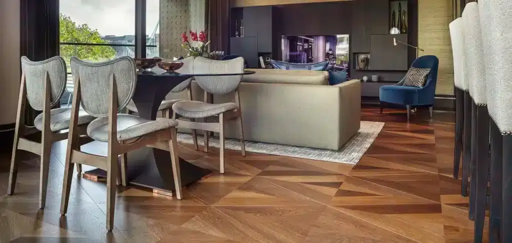 A modern dining and living area featuring bespoke wood flooring with a unique light diamond pattern. The space includes a stylish dining table with cushioned chairs, a comfortable beige sofa, and a blue armchair. Large windows offer a view of a green landscape, adding natural light to the sophisticated interior design.