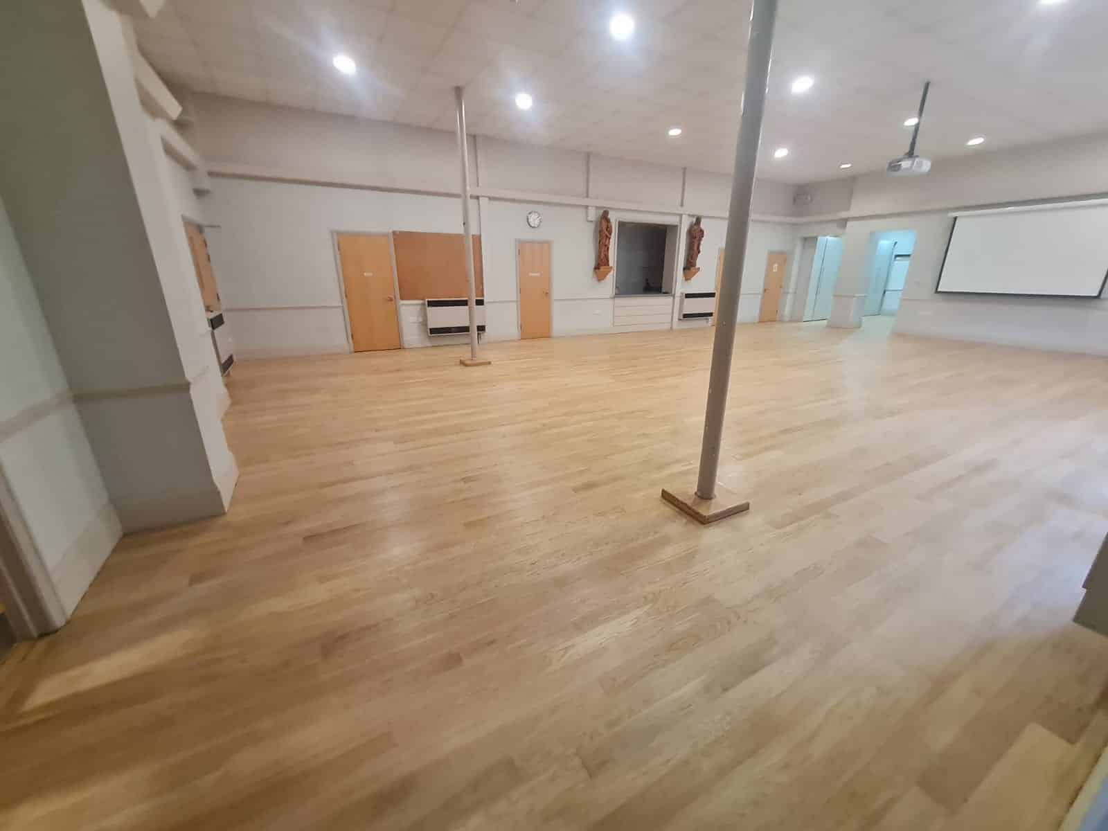 A large room with a freshly refinished wooden floor, completed by Mr Sander®. The floor has a smooth, polished appearance. The room features high ceilings, several doors, a clock, and a projector screen. The space is well-lit, highlighting the pristine condition of the floor.