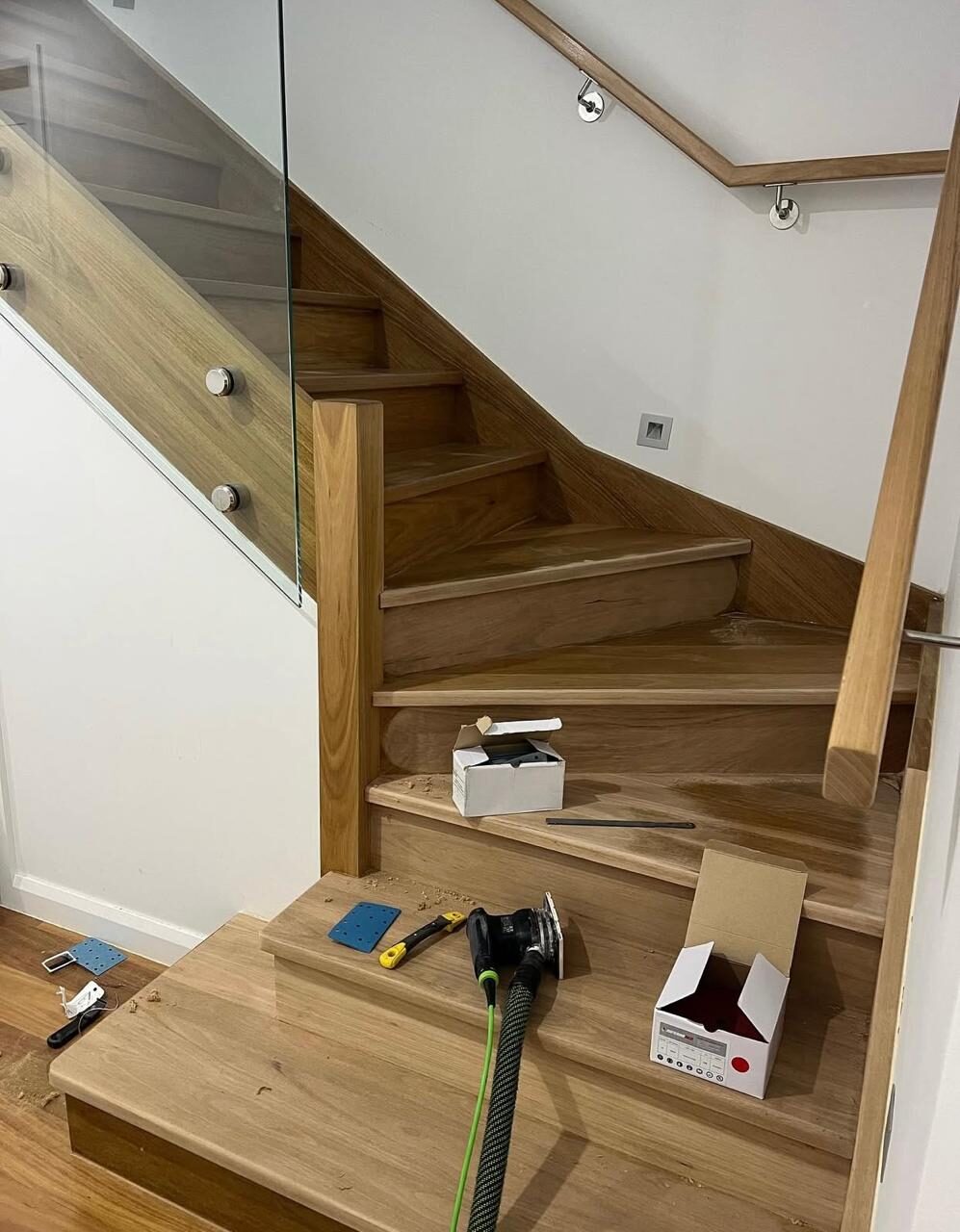 Professional tools and sanders laid out on a modern solid oak staircase with glass balustrades during the process of sanding stair treads.