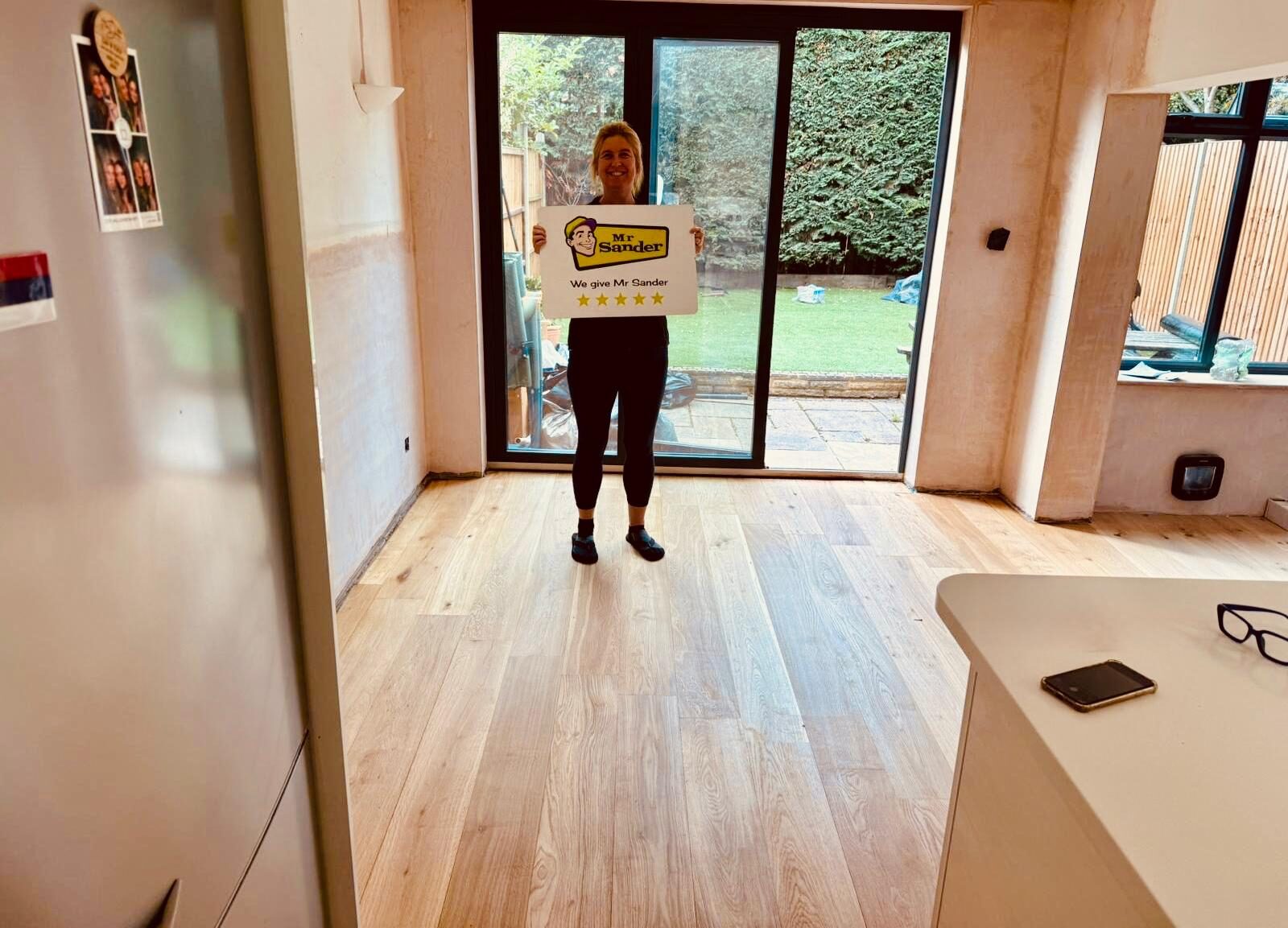 Happy client standing on newly restored oak floor holding Mr Sander sign—after Repairing Scratches on Hardwood Floors in a bright kitchen-diner