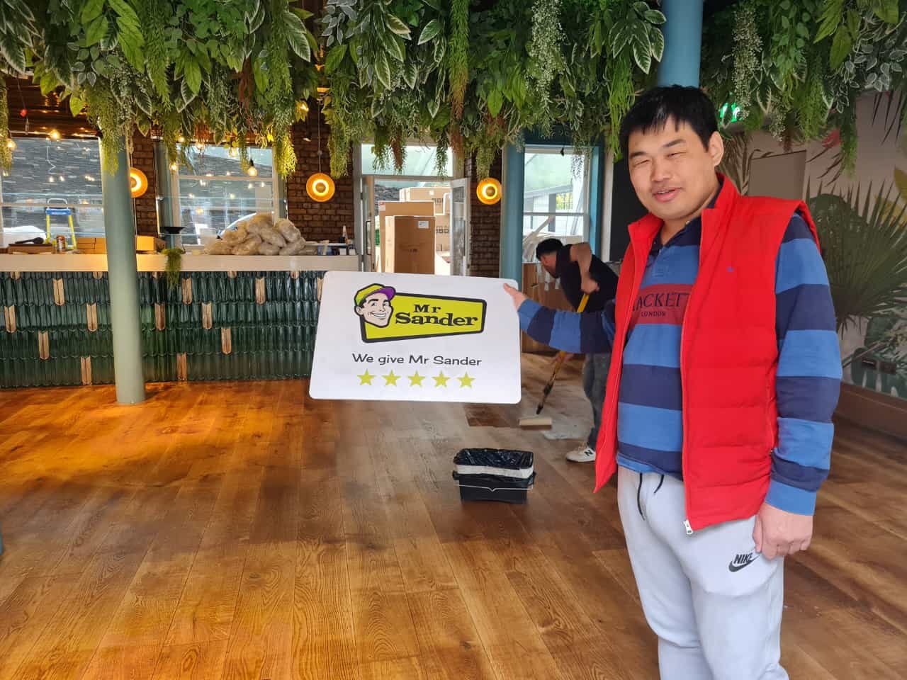 Happy customer holding a Mr Sander review sign in a UK restaurant after professional floor sanding, with newly restored wooden floors.
