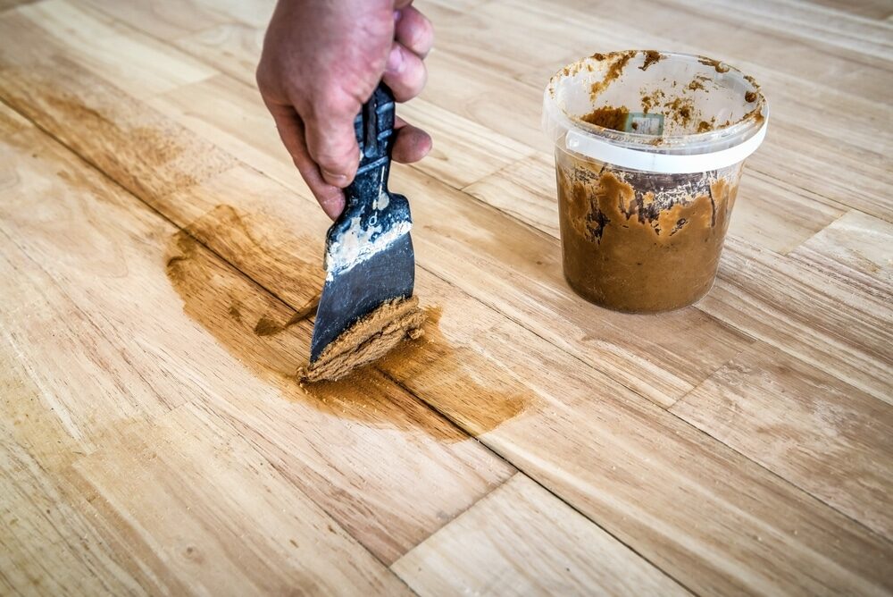 Restore Engineered Wood Floors – close-up of engineered oak plank flooring being repaired with wood filler applied into gaps and cracks using a scraper.