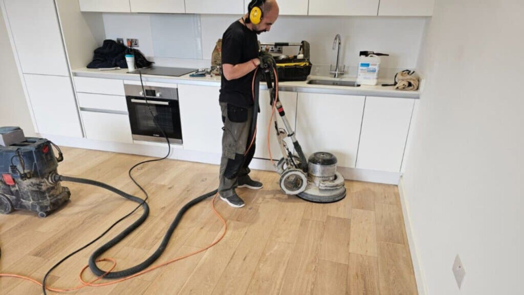 Professional technician using a dust-free sanding machine on a light oak kitchen floor before French polishing to restore pet-stained floors.