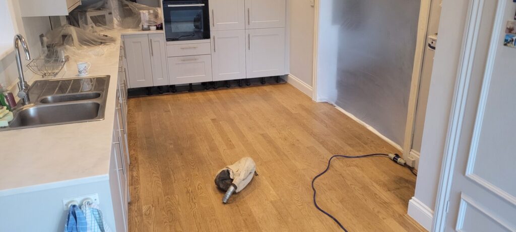 Sanded solid oak plank kitchen floor prepared for French polishing, showing dust-extraction bag and masked cabinets—next step to Restore Pet-Stained Floors.