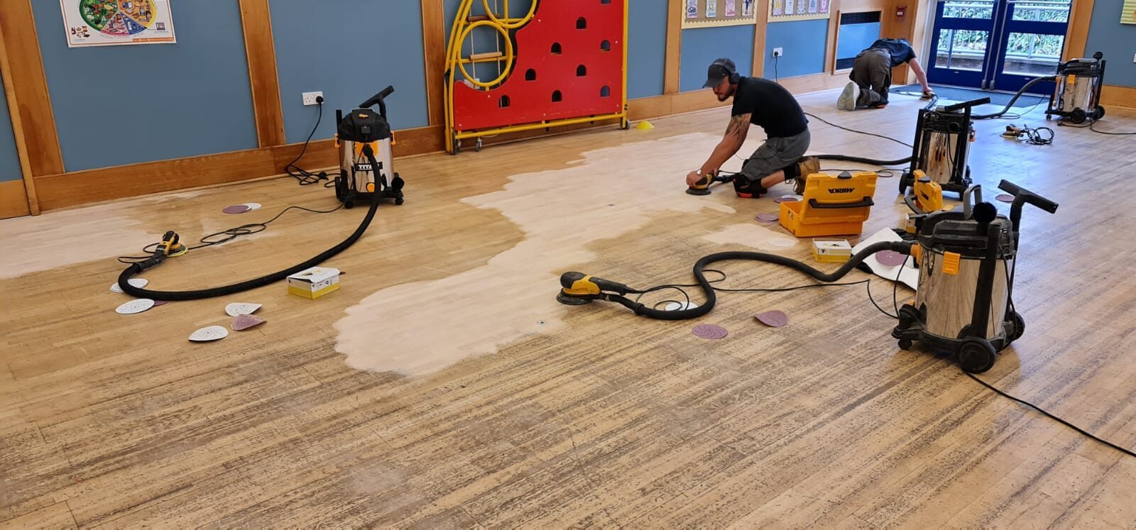 Restoring Floors in Schools – technicians sanding a school hall’s maple strip wood floor with dust-extraction equipment, preparing for child-safe, low-VOC finishes.
