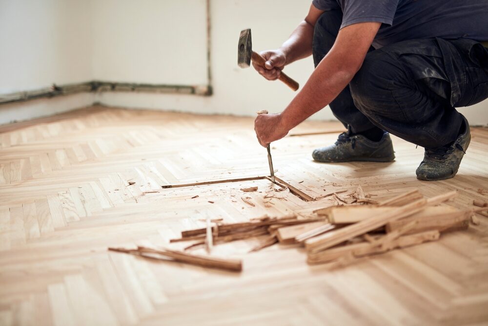 Restoring Parquet Flooring – tradesperson removing damaged oak herringbone parquet blocks with a hammer and chisel before repairs and sanding.