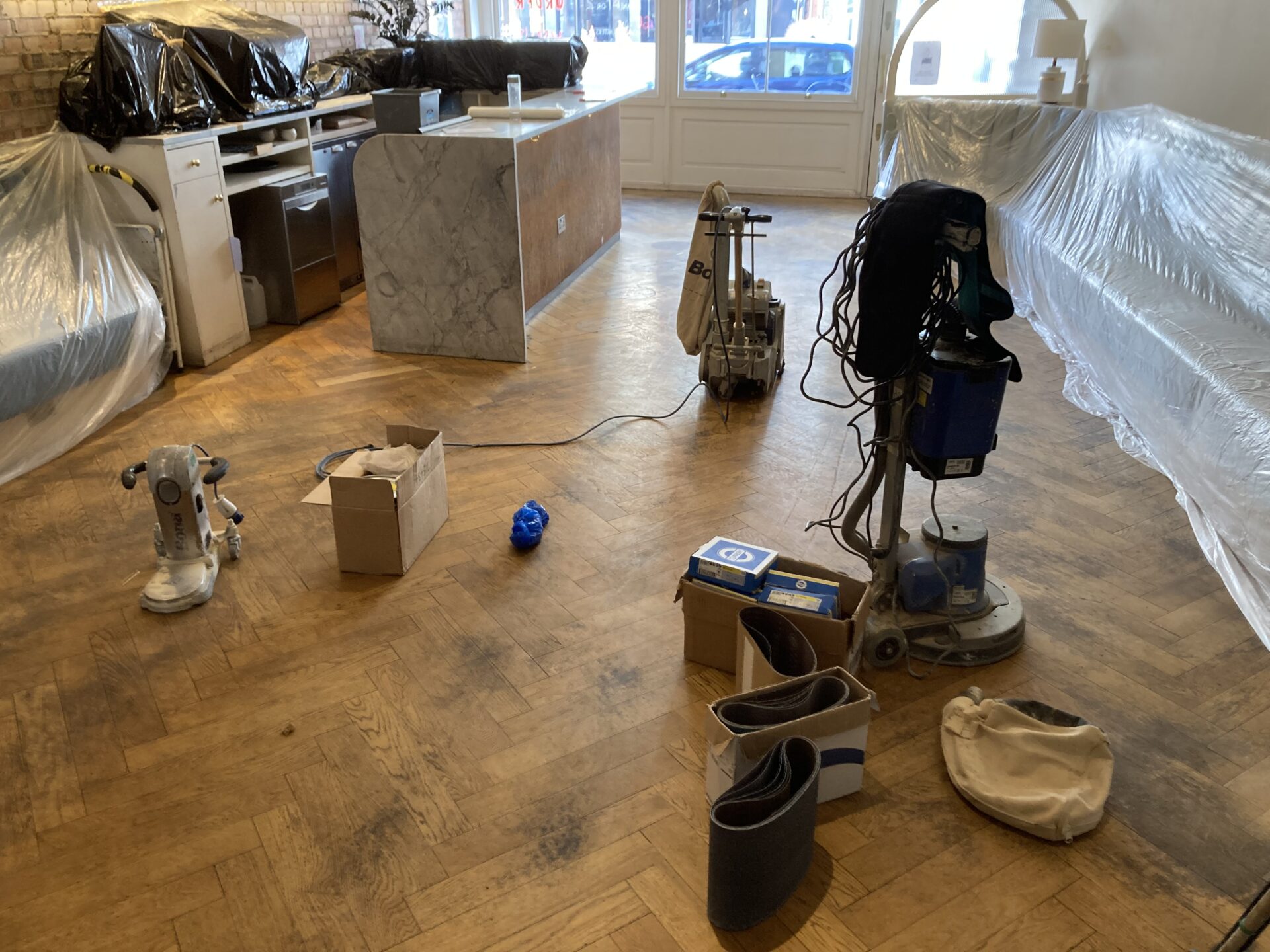 Restoring Parquet Flooring setup on a worn herringbone oak parquet floor—professional belt sander, edger and rotary buffer with abrasives laid out, furniture and seating protected for dust control.