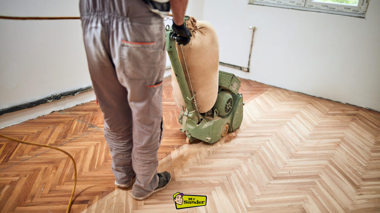 Restoring Parquet Flooring in progress with a belt sander on herringbone oak parquet—half sanded to bare wood, half with the old finish visible.