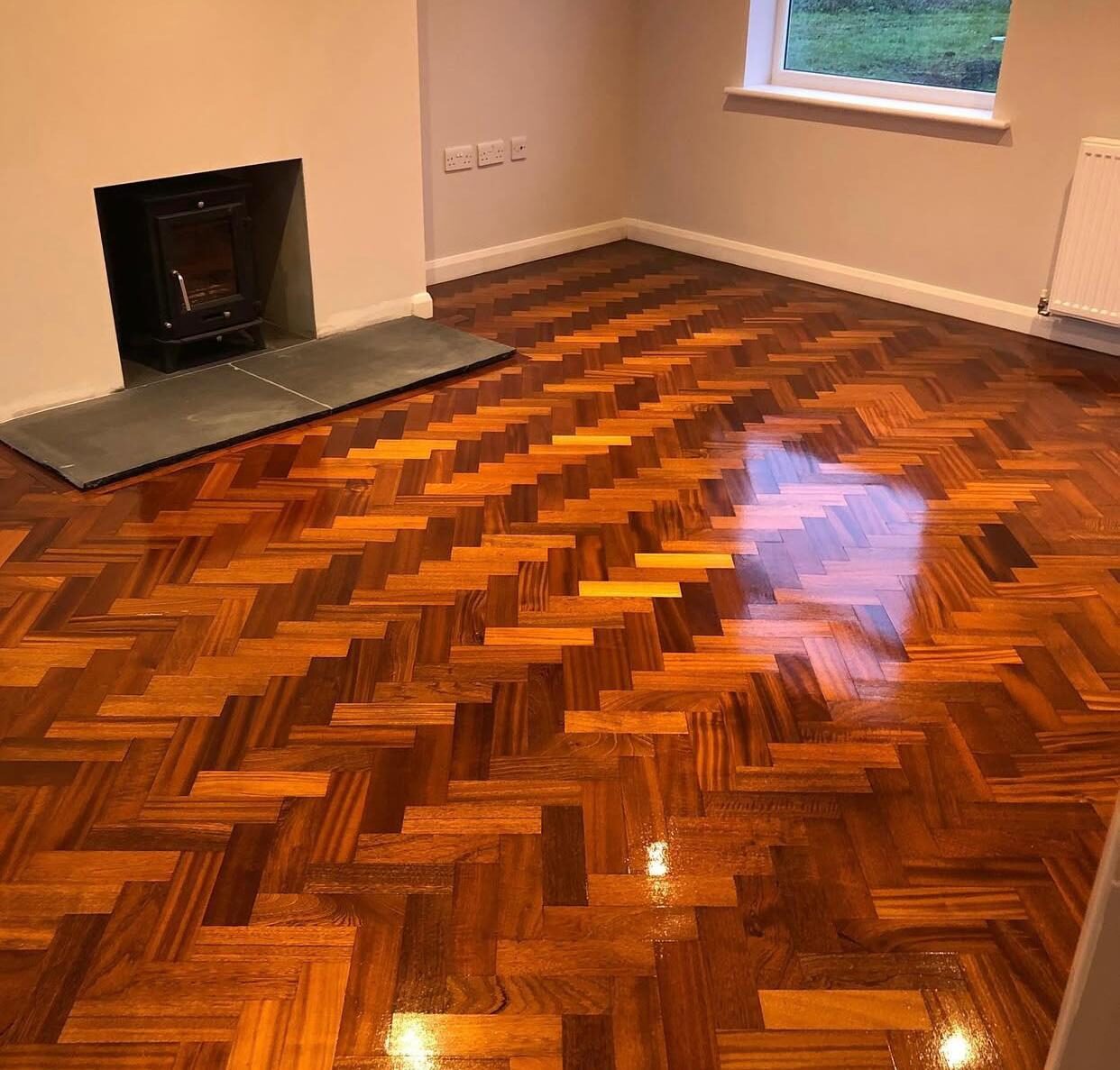 Restoring Parquet Flooring on a classic herringbone solid hardwood parquet—repaired, finely sanded, stained warm amber and sealed to a mirror-gloss finish beside a stove hearth.