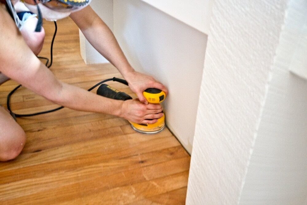Person using a yellow orbital sander on a wooden floor next to a white wall.