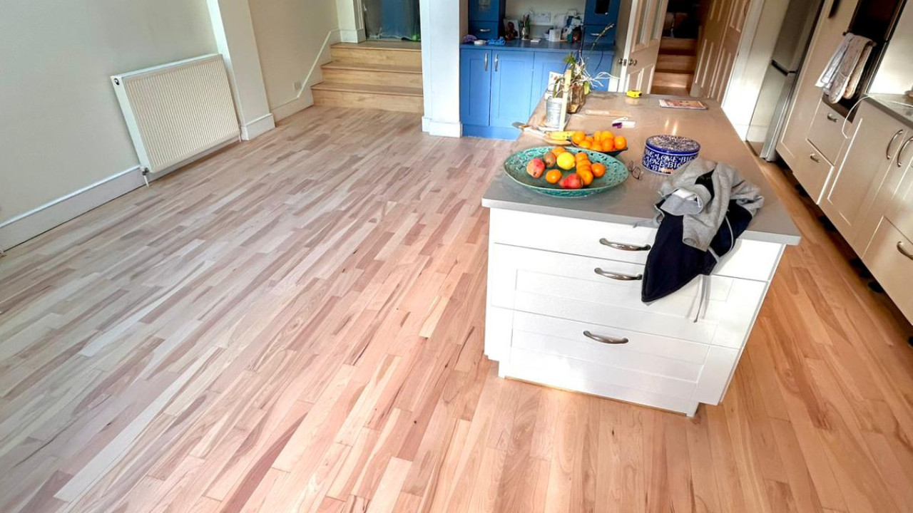 A wide view of a kitchen and dining area during the process of sanding and refinishing oak floors, showing the raw light wood before the final finish.