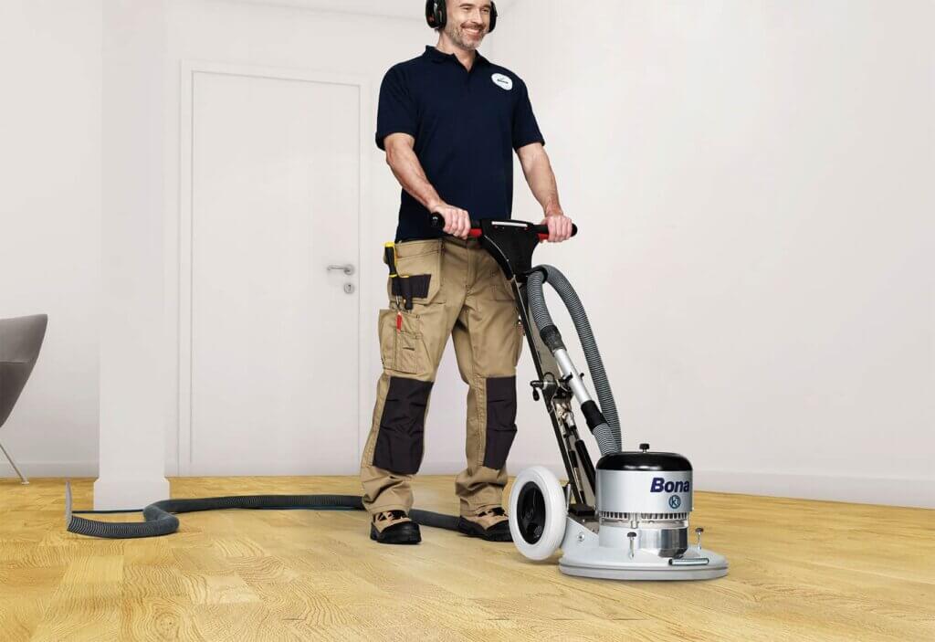 A Mr Sander® professional is sanding an oak floor in a living room in Welling, UK, using a Bona FlexiSand 1.9 (AMO310002) buffer. The machine features a 1.9 kW motor, 230 V, 50 Hz, with a 407 mm disc diameter, 147 rpm speed, 50 kg weight, and safety devices including a 16 A overload switch and thermal protection.