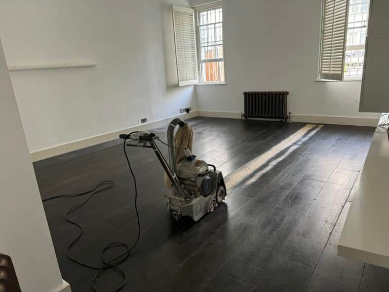 A spacious room with dark-stained wooden floors being sanded using professional floor sanding equipment, with natural light streaming through shuttered windows.