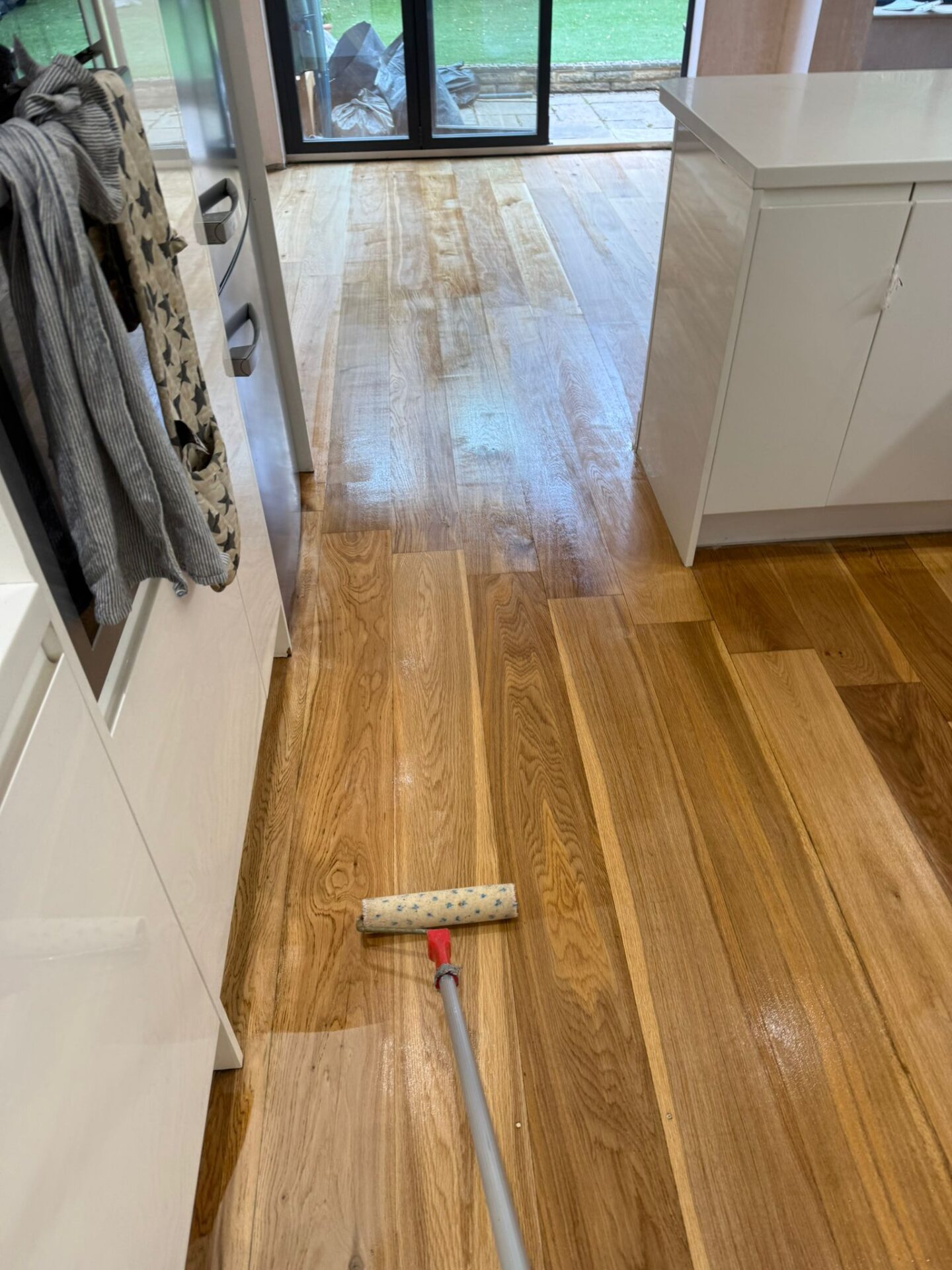 Applying finish to oak kitchen floor with a roller after sanding—midway through sanding, staining and finishing wood floors, near patio doors.