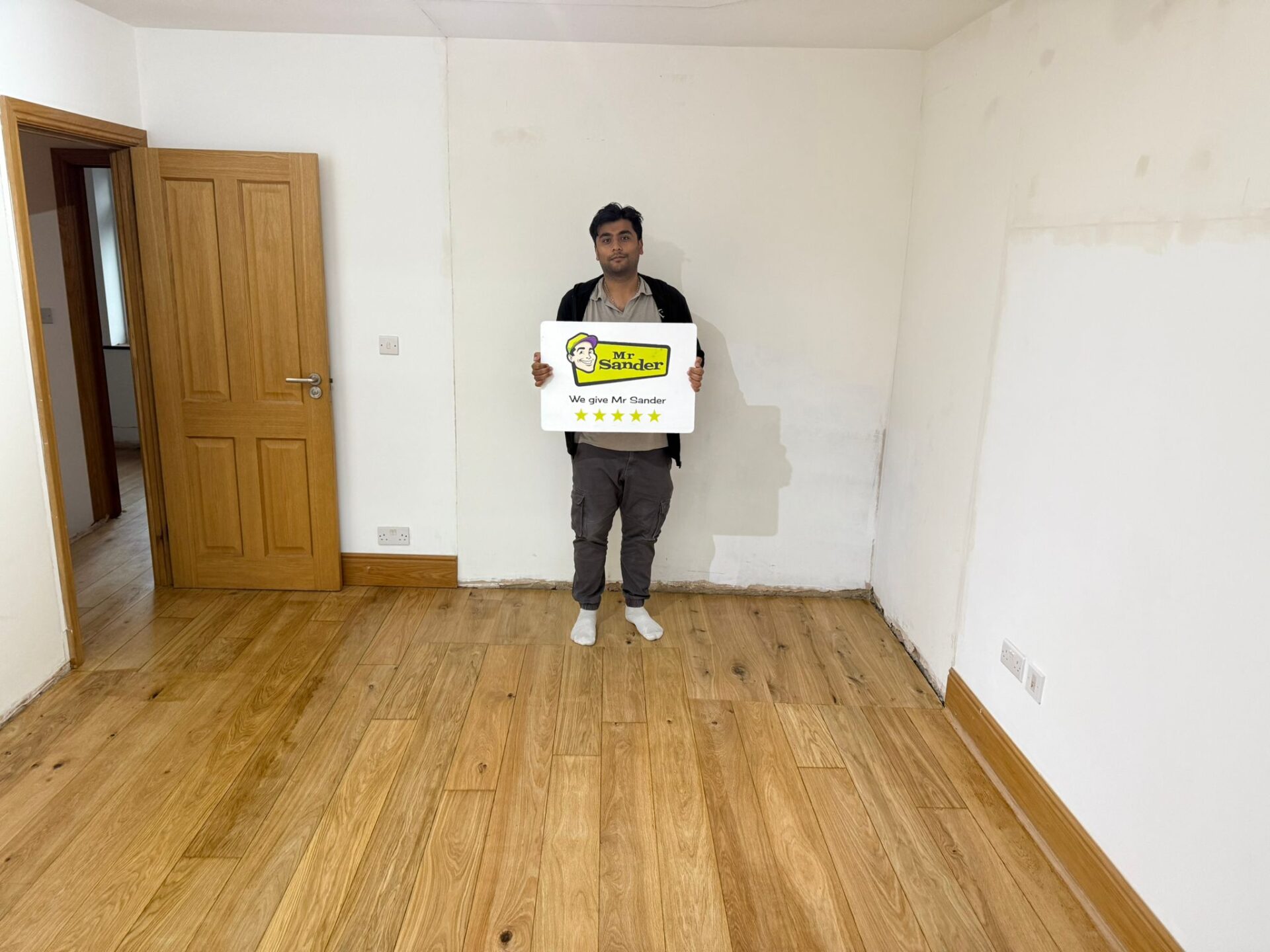 Sanding Wood Floors Successfully — satisfied customer holding a Mr Sander® five-star sign above a freshly restored oak plank floor with a clear lacquer finish.