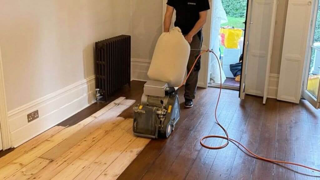 A man from Mr Sander® is sanding a wooden floor in a living room using a Frank Cobra Belt Sander. The machine, weighing 78 kg, features a powerful 2500W motor, operates at 2100 rpm, and uses a 200 x 750 mm endless belt. The Cobra offers 99.99% dust extraction through its integrated DCS system. It includes features like a quick-change belt system, Featherlite clutch control for precision sanding, and an automatic belt adjustment control. The machine is suitable for professional sanding and can be dismantled for easy transportation.