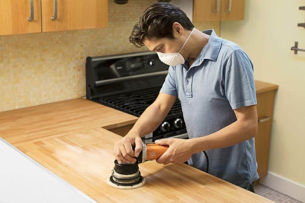 Man sanding a wooden butcher block countertop in a kitchen while wearing a dust mask.