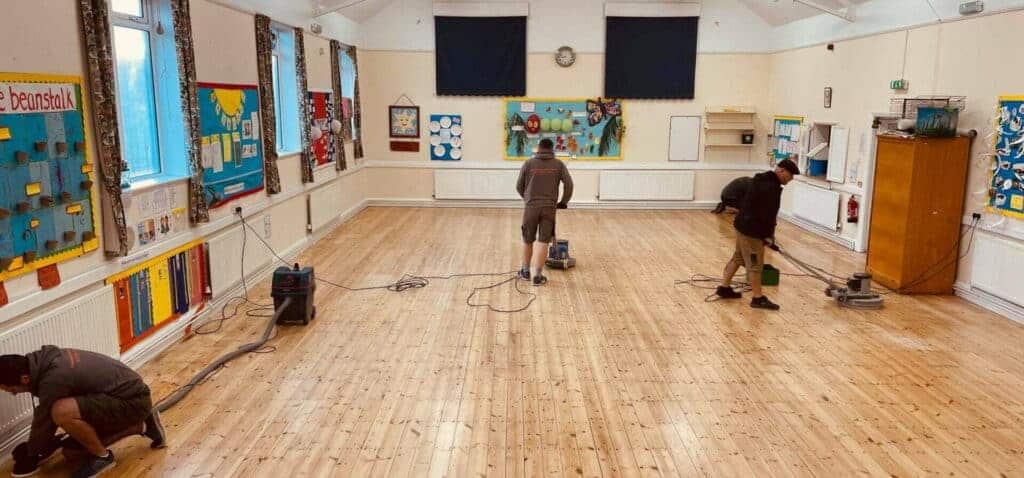 Four professionals from Mr Sander® working on a school pine floor in Childs Hill, UK. They are sanding, repairing, buffing, and oiling the floor using the Bona FlexiSand 1.5 (AMO310001). This machine features a 1.5 kW single-phase motor, 230V voltage, 50 Hz frequency, and a 407mm disc diameter. It includes safety devices like a 10A overload switch and thermal protection. The machine operates at 147 rpm unloaded, weighs 45kg, and has dimensions of 1170mm height, 490mm width, and 640mm depth.
