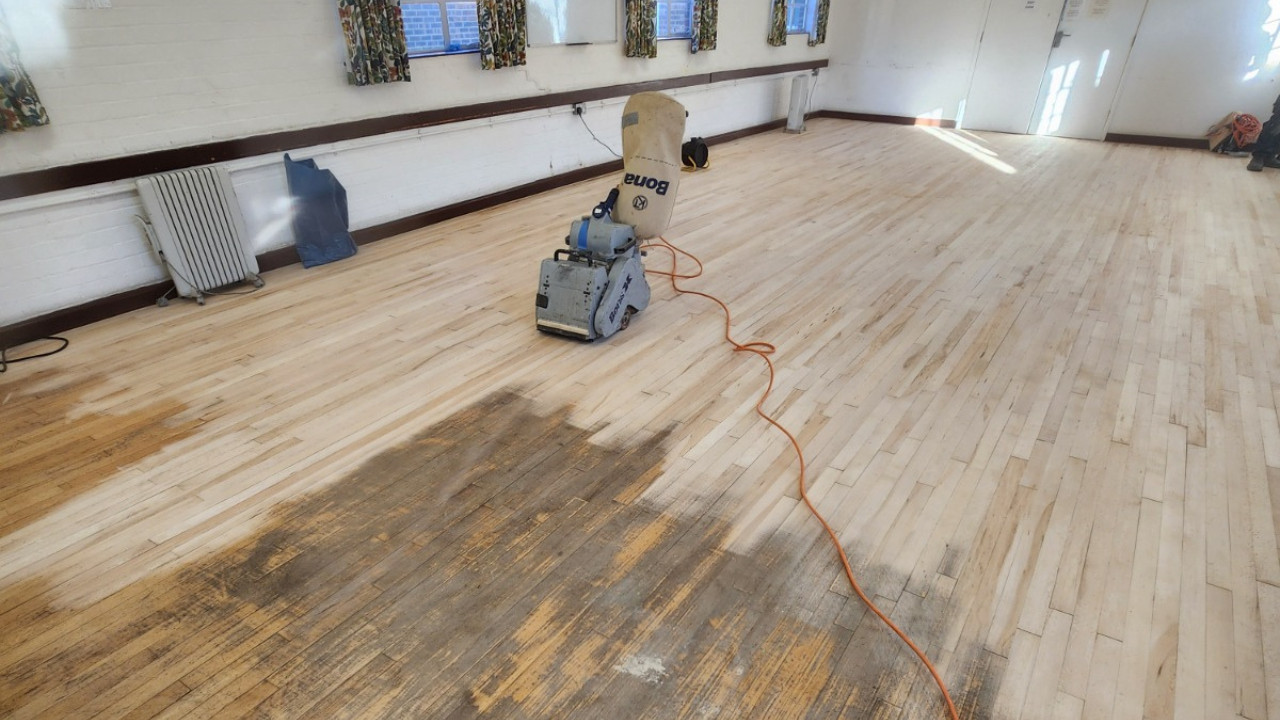 A Bona belt sander being used on a solid hardwood Junckers beech floor during a school hall maintenance project to remove old finish and scratches.