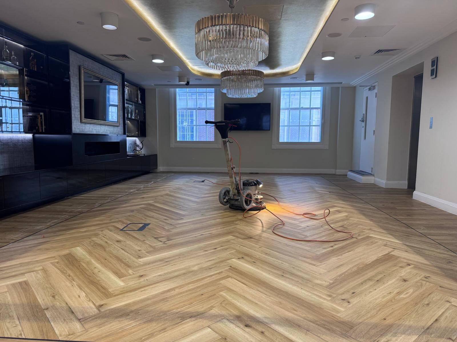 A professional sanding machine screening a large herringbone wood floor in a modern interior, with lighting highlighting the use of the right screen grit for smooth, even finish preparation.