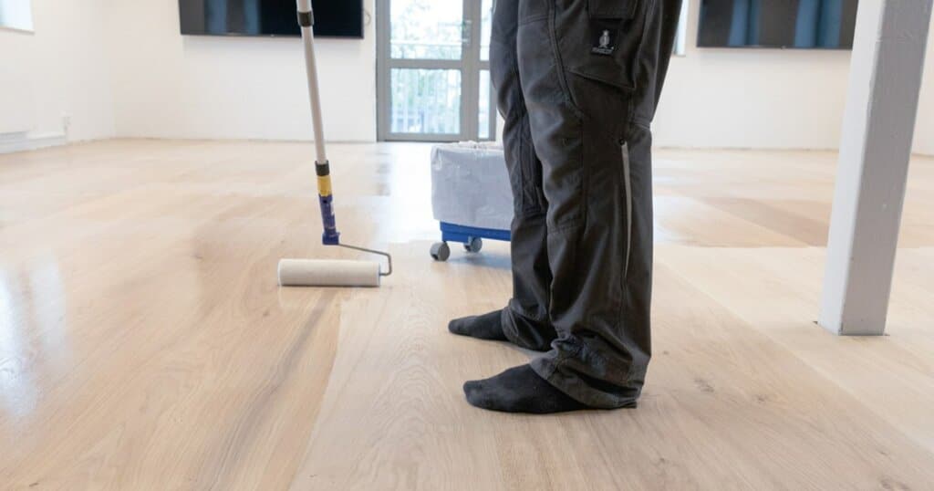 A man from Mr Sander® is sanding and refinishing a wooden floor in a UK living room using a professional Bona Belt sander (model AMO110000.3) with a 2.2 kW motor, 230 V, 50 Hz, and a drum width of 200 & 250 mm. The floor is being refinished with a roller applicator. The room has a minimalistic design with a large window and wall-mounted TV.