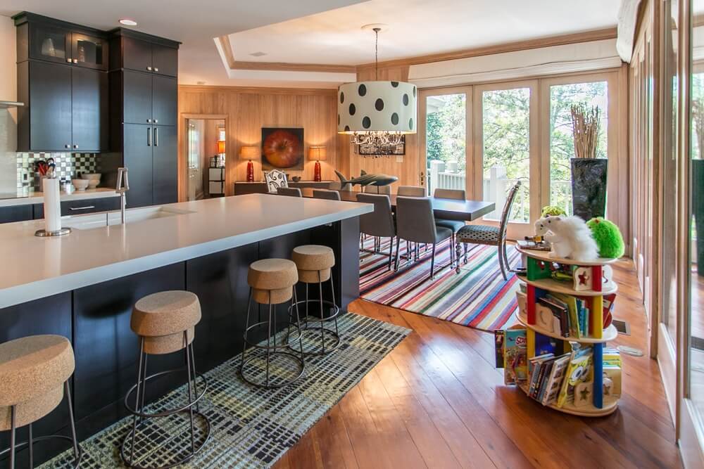 A spacious, modern kitchen and dining area featuring beautifully refinished warm-toned hardwood floors. The room includes a large kitchen island with bar stools, a colorful striped rug under the dining table, and expansive windows that let in natural light. The professional sanding and refinishing of the hardwood floor by Mr Sander® adds a luxurious, polished touch to the space.