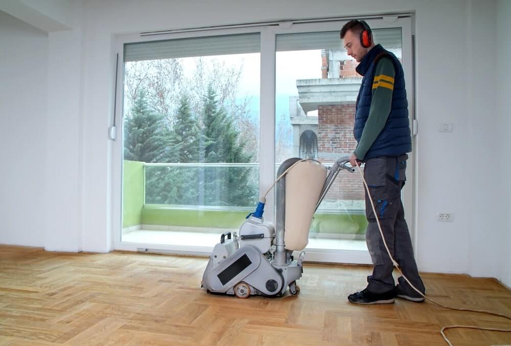 A man from Mr Sander® sanding a parquet floor in a living room in East Sheen, UK using a Bona Belt (AMO110000.3) belt sander. The machine operates at 2.2 kW, 230 V, 50 Hz, with a sanding belt size of 250x750 mm. Drum widths available are 200 mm and 250 mm. The sander weighs 74 kg.