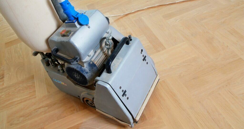 A man from Mr Sander® is sanding a parquet floor in a living room in Brondesbury, UK, using a Bona Belt sander (AMO110000.3) with a motor power of 2.2 kW, operating at 230 V and 50 Hz, and a drum width of 250 mm. The sander weighs 74 kg and uses a 250x750 mm sanding belt.