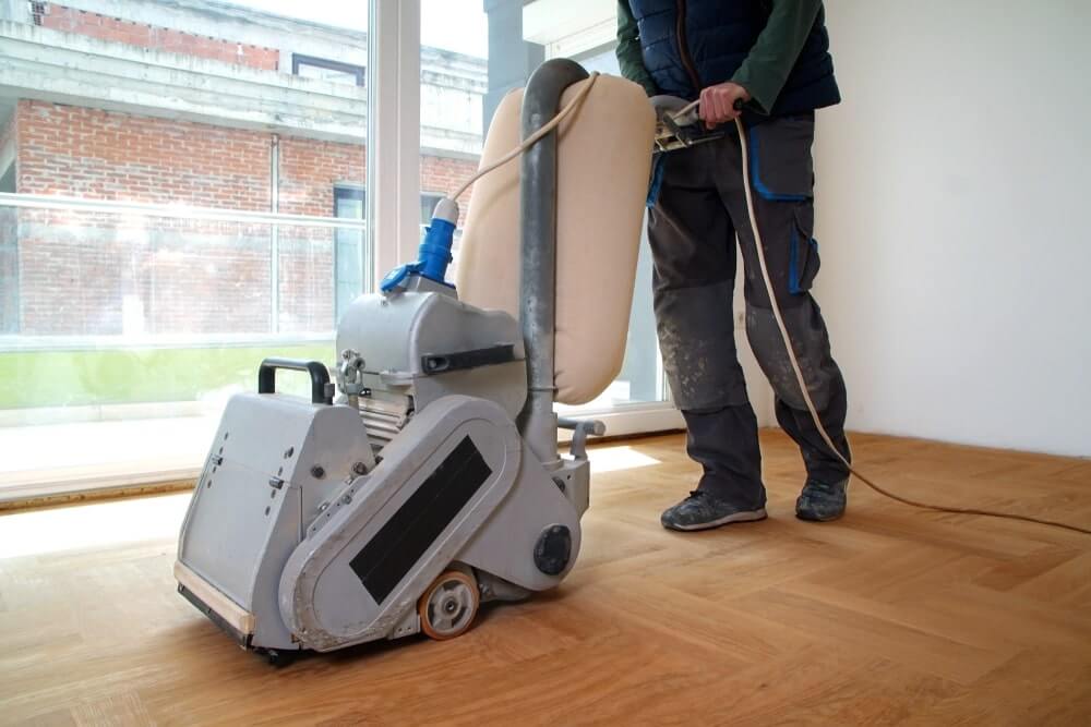 A professional from Mr Sander® using a Bona Belt sander (model AMO110000.3) to sand a floor in a living area in the UK. The belt sander has an effect of 2.2 kW, operates at 230 V and 50 Hz, and measures 250x750 mm. The drum width is 200 & 250 mm, and the sanding belt size is 200 x 750 mm or 250 x 750 mm. The motor power is 2.2 kW, and the total weight of the machine is 74 kg.