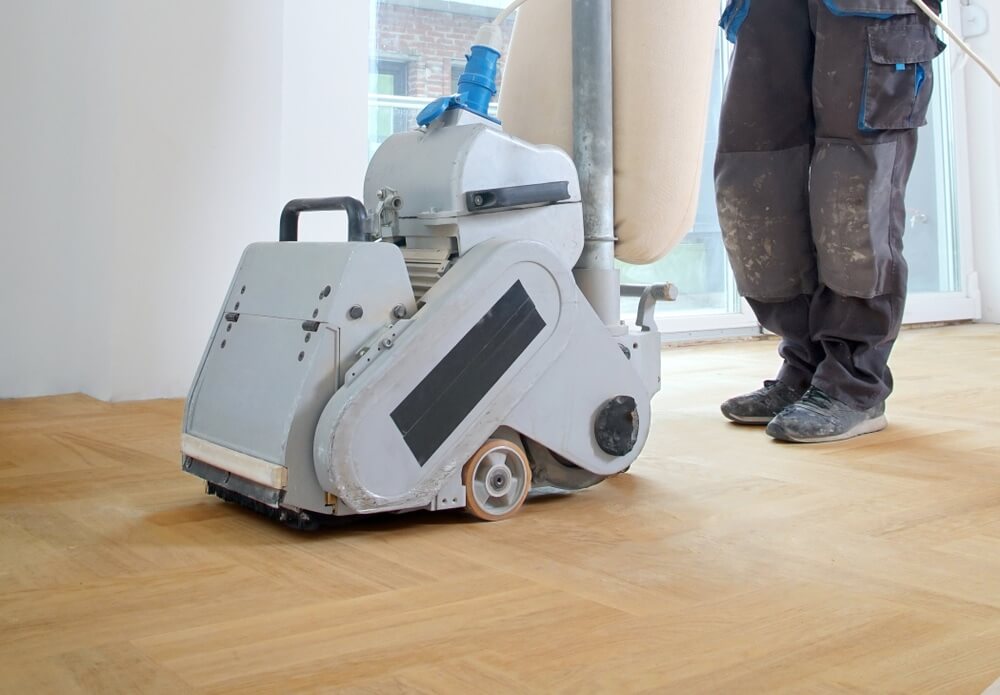 A professional from Mr Sander® using a Bona Belt sander (model AMO110000.3) to sand a floor in a living area in Belsize Park, UK. The belt sander has an effect of 2.2 kW, operates at 230 V and 50 Hz, and measures 250x750 mm. The drum width is 200 & 250 mm, and the sanding belt size is 200 x 750 mm or 250 x 750 mm. The motor power is 2.2 kW, and the total weight of the machine is 74 kg.