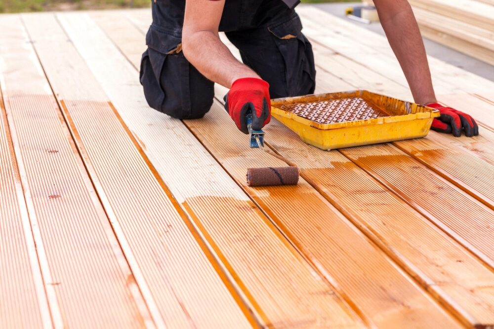 A person kneeling and applying stain to a wooden deck using a paint roller.