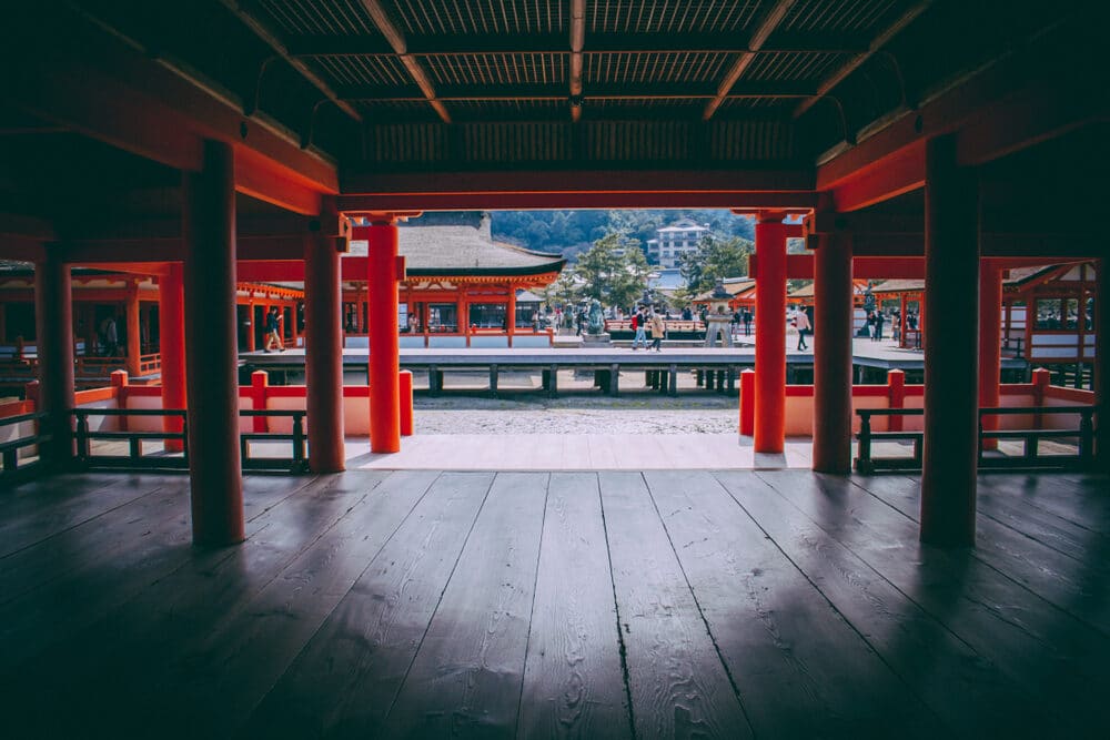 View from inside a traditional Japanese temple with red pillars and a wooden floor, looking out towards a courtyard.