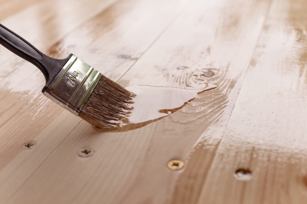 A close-up of a paintbrush applying a clear varnish or finish to a wooden floor, highlighting the smooth, shiny surface of the freshly coated wood.