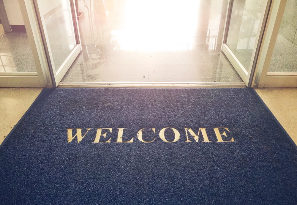 A welcome mat placed at the entrance of a building with open glass doors. The mat is dark blue with the word "WELCOME" printed in large white letters. The interior beyond the doors features a glossy floor, suggesting a clean and polished environment.