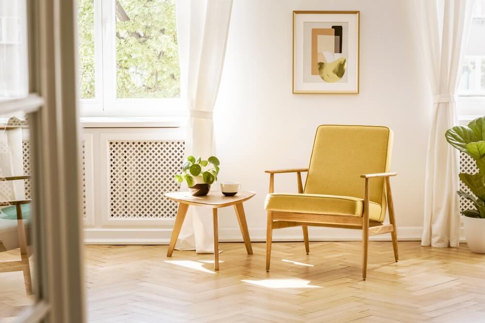 A bright and cozy living room in Dalston, UK, featuring a freshly sanded and refinished herringbone wooden floor, a yellow armchair, a small wooden table with a plant, and a window letting in natural light. Showcasing the expert floor sanding and restoration services by Mr Sander®.