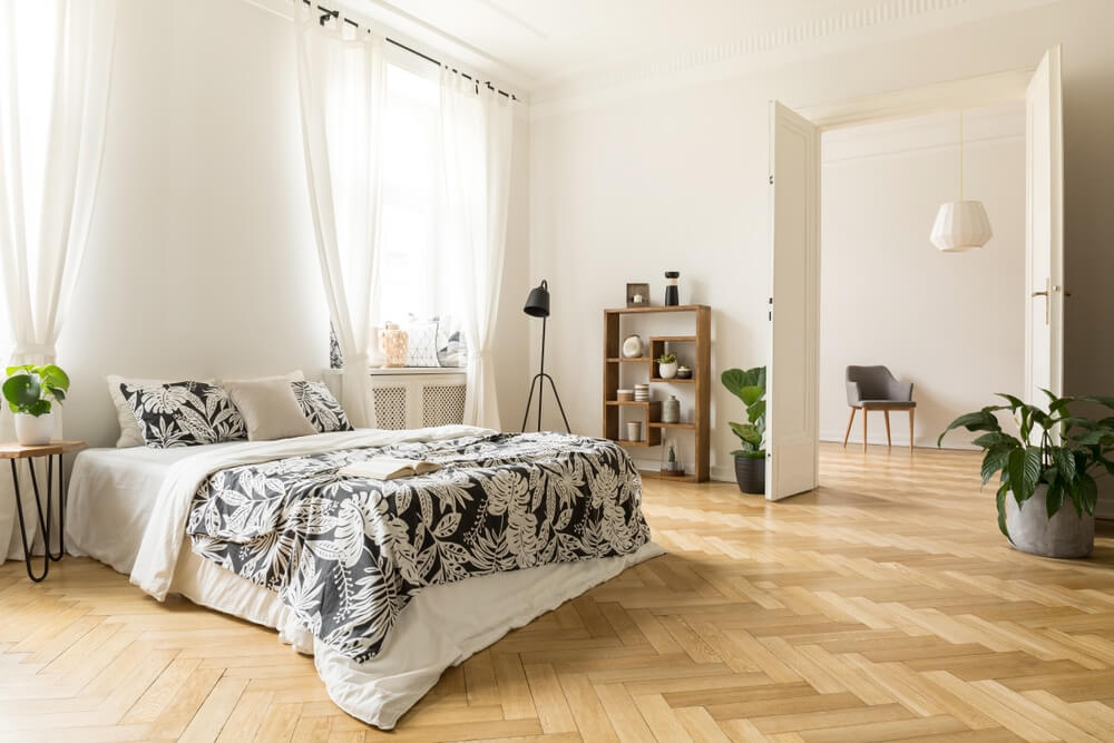 A beautifully restored parquet floor enhances the elegance of a bright, modern bedroom. The room features a bed with black and white floral bedding, light curtains, indoor plants, and minimalist decor, showcasing the timeless appeal and sophistication achieved through professional parquet flooring restoration.