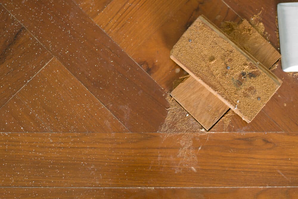A close-up image of a wooden floor being cleaned with a sponge, showing the contrast between the cleaned and dirty areas