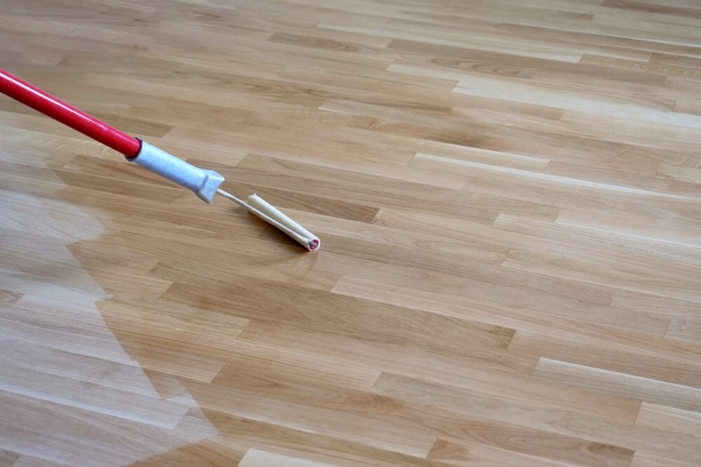 A roller is being used to apply a clear finish to a freshly sanded wooden floor, highlighting the even application and the wood's natural grain. The floor has a smooth and polished appearance, showcasing the professional refinishing work by Mr Sander®.