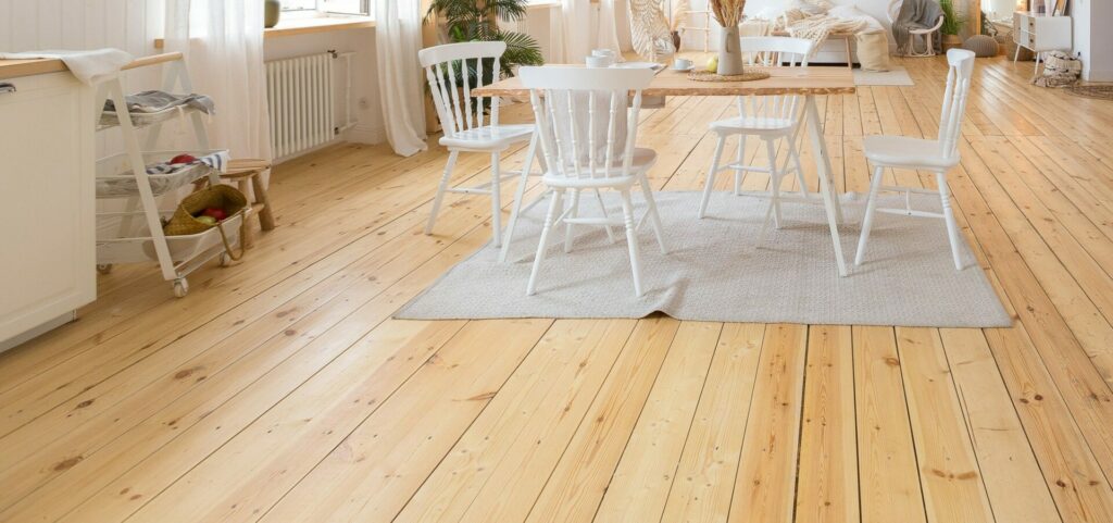 A bright and airy dining area with wide, light-coloured wooden floorboards that enhance the natural, cosy atmosphere of the room. A simple wooden table with white chairs is placed on a light rug, while natural light floods in through large windows, highlighting the clean and polished finish of the floor.