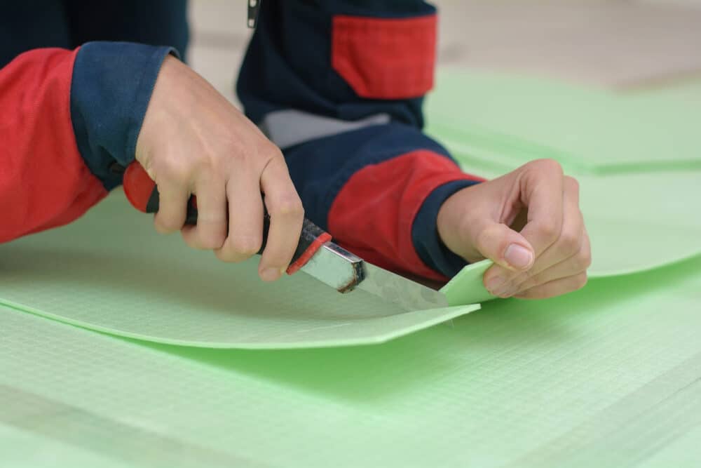 Close-up of hands cutting green floor underlayment with a utility knife.