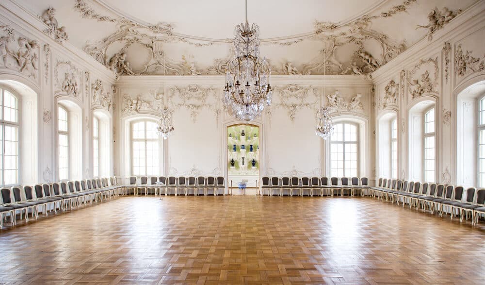 Opulent baroque hall with ornate stucco ceiling, crystal chandeliers, and rows of blue upholstered chairs on parquet flooring.