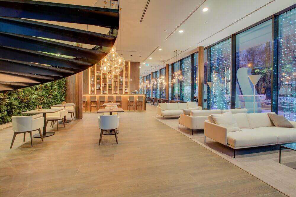A modern, luxurious interior space with wooden floors that have been sanded and refinished by Mr Sander®. The area features a spiral staircase, white sofas, and tables with chairs. Large floor-to-ceiling windows provide a view of the outside, while stylish pendant lights and a green wall add to the elegant decor.